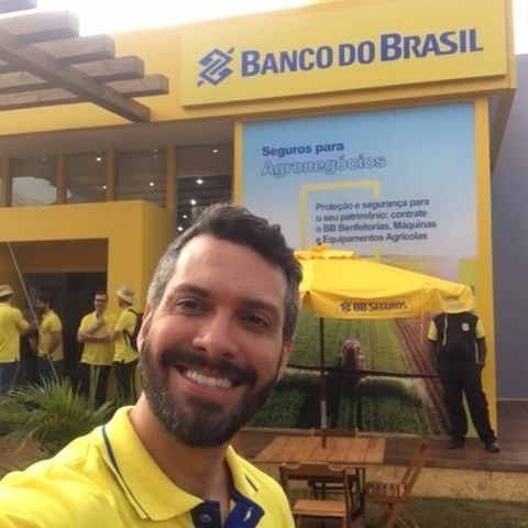 Paulo Hora fazendo selfie, vestindo camiseta amarela da BB Seguros. Ao fundo, uma estrutura com o logo do Banco do Brasil e, abaixo dele, um banner divulgando seguros para o agronegócio.